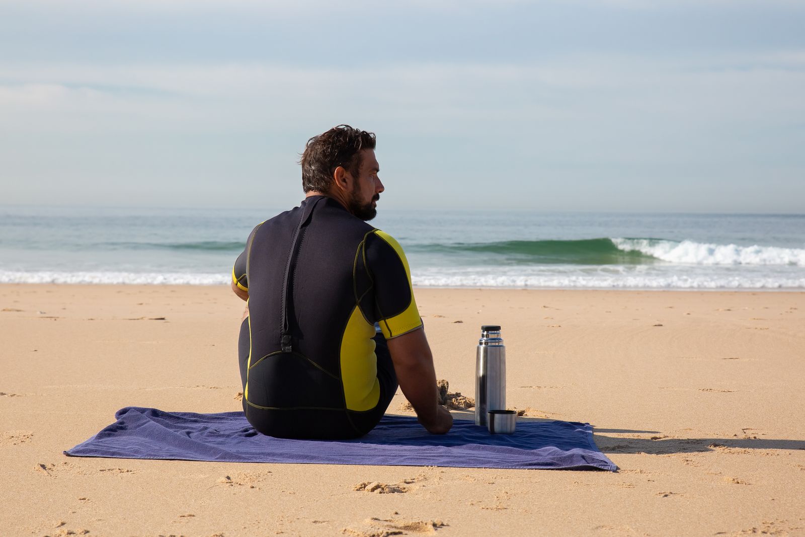 A swimmer in a wetsuit sitting on the shore with a flask, warming up after a cold swim